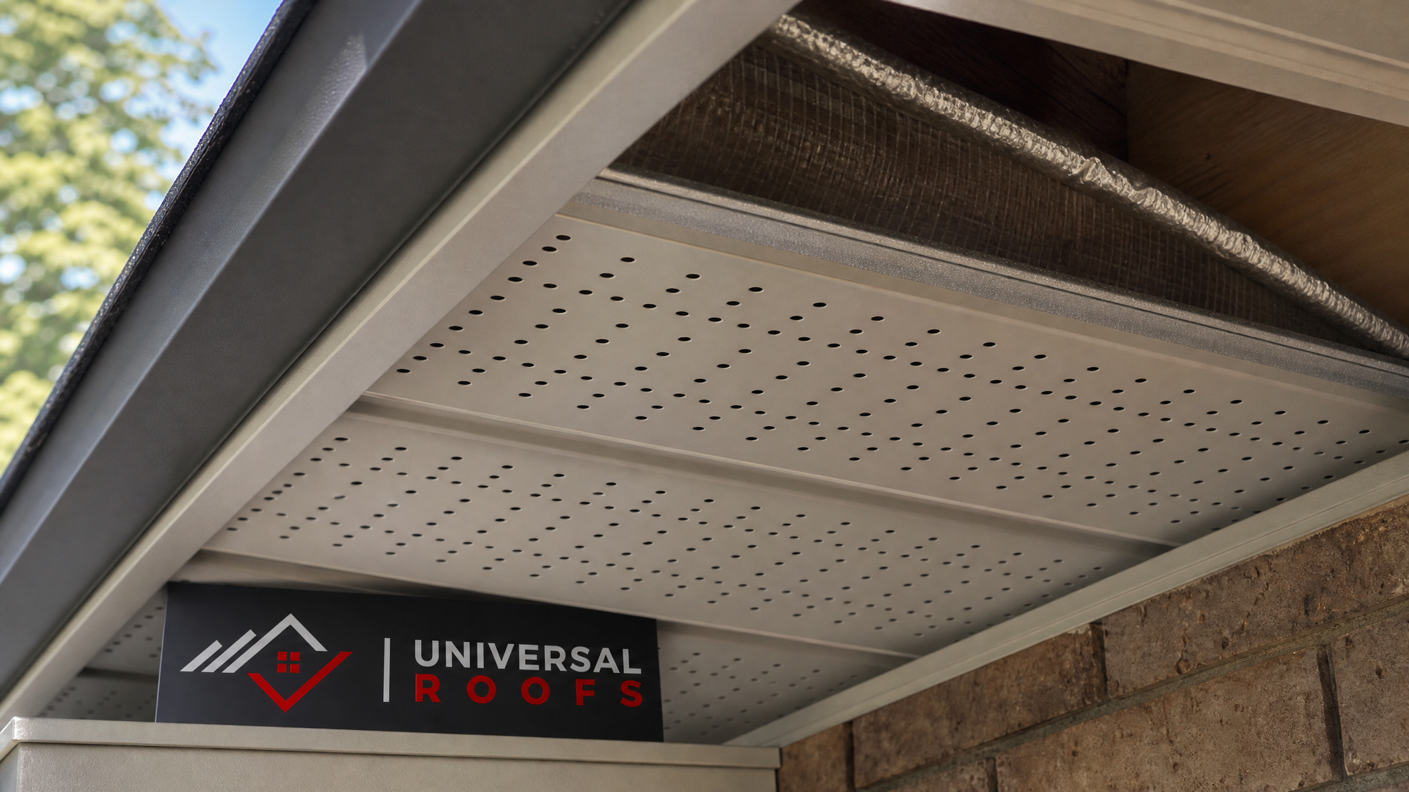 Close-up of vented aluminum soffit panel installed correctly with insulation baffle visible in the rafter bay, branded sign placard beside it
