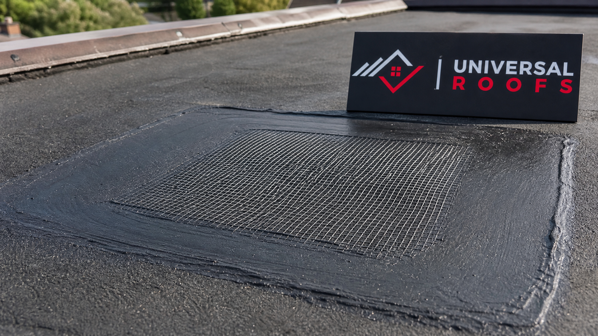 Close-up detail of a reinforced flat roof patch seam showing fibreglass mesh embedded between two layers of sealant, with a small branded sign placard beside it