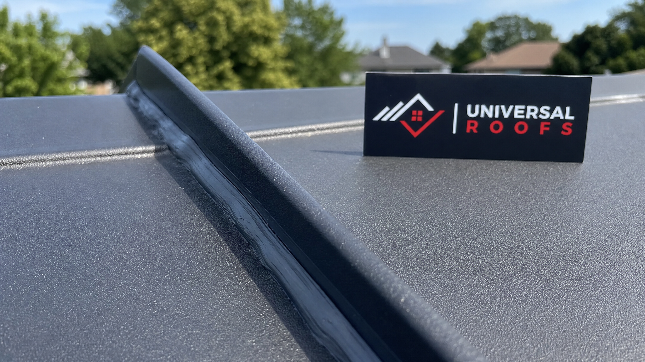 Close-up of a standing seam metal roof fold with fresh butyl sealant bead visible and a Universal Roofs branded sign placard nearby