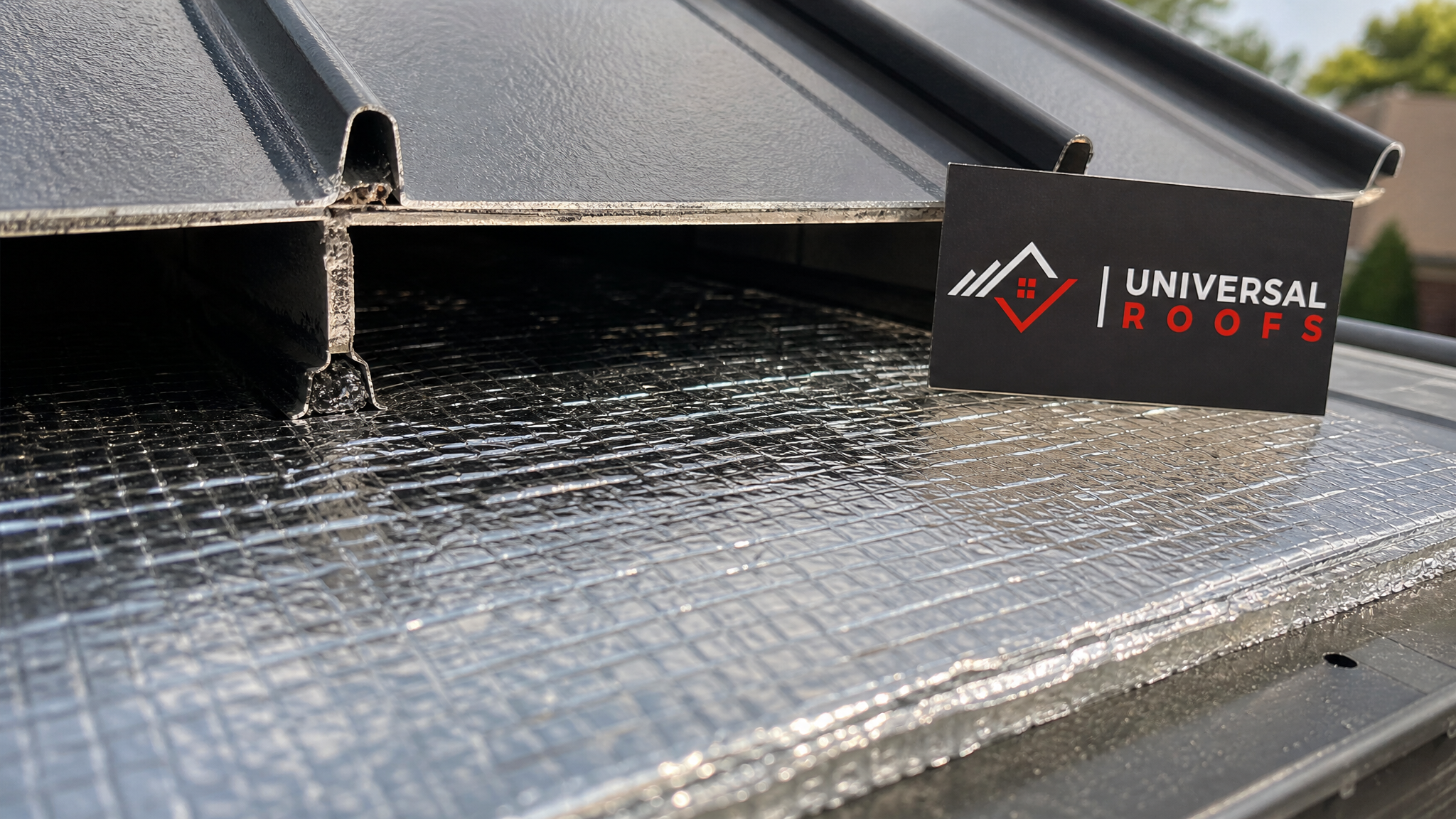 Close-up of radiant barrier foil insulation and vent channel detail under a metal roof panel with a Universal Roofs branded sign placard