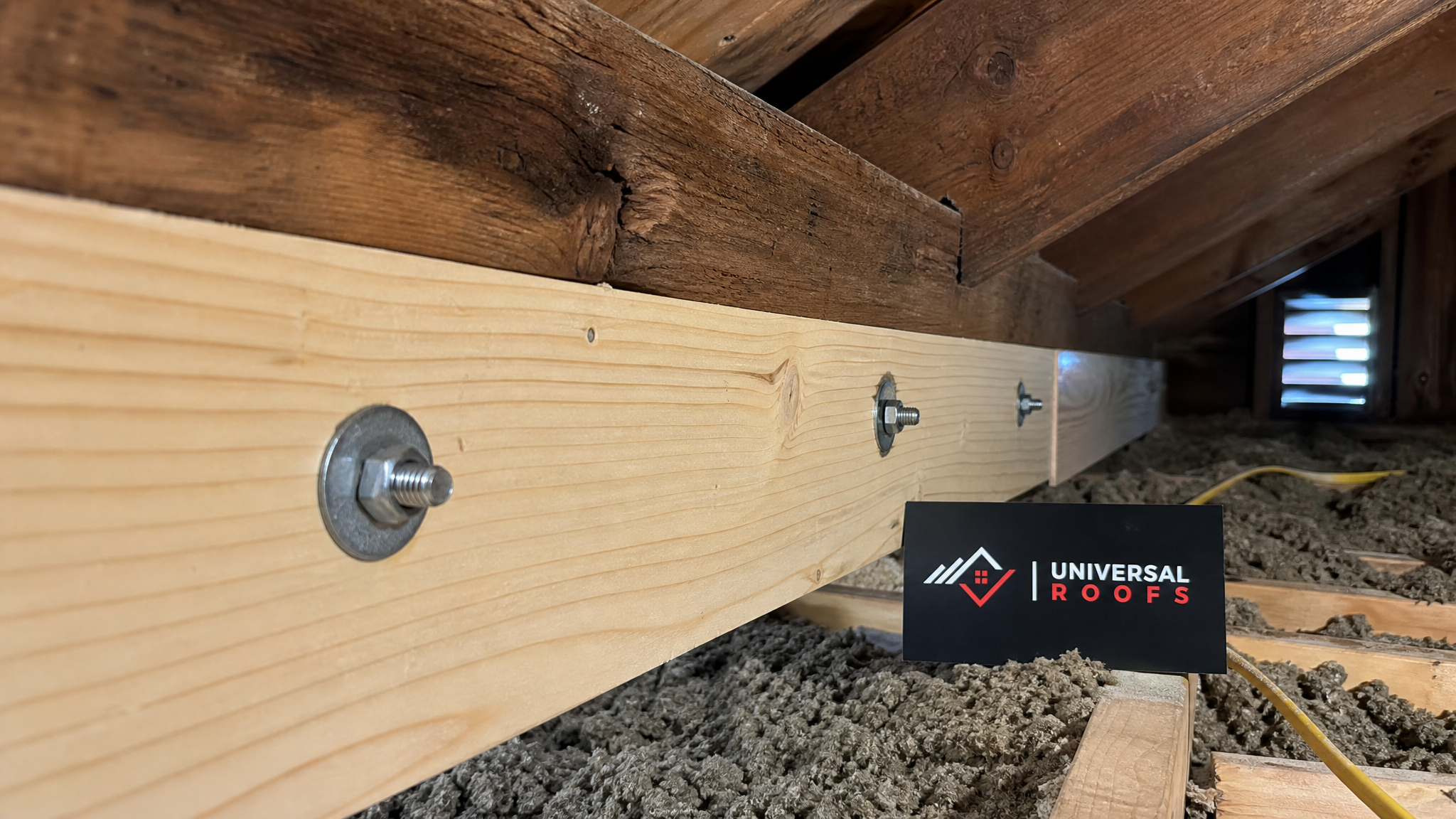 Close-up detail of a reinforced wood rafter joint with a Universal Roofs branded sign placard nearby, showing sistered lumber bracing technique