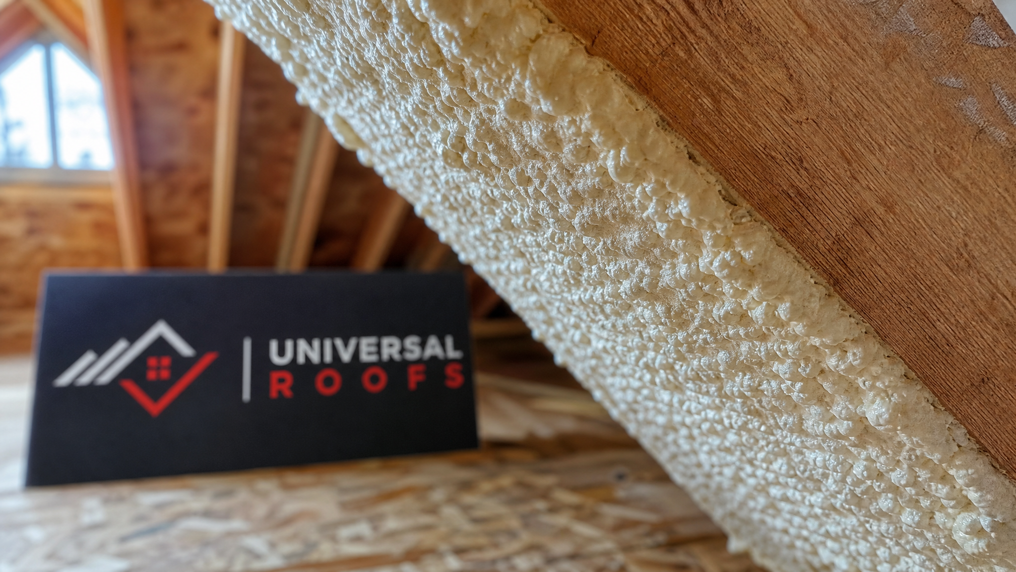 Close-up detail of cured closed-cell spray foam insulation showing its dense, rigid cell structure bonded tightly to wood roof rafters, Universal Roofs branded sign placard visible nearby, natural daylight