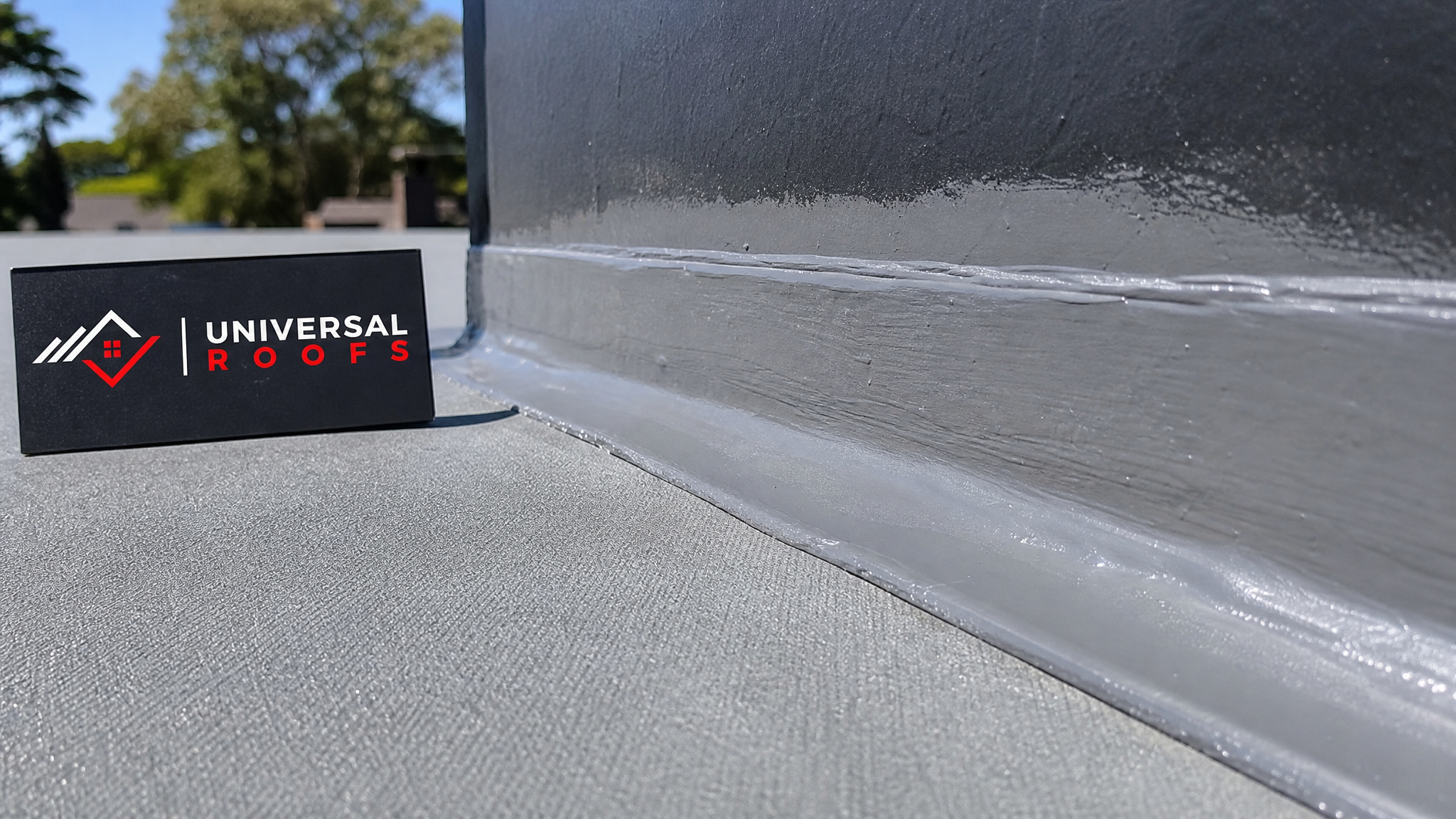 Close-up of a newly sealed roof seam and flashing joint on a flat roof with a Universal Roofs branded sign placard nearby