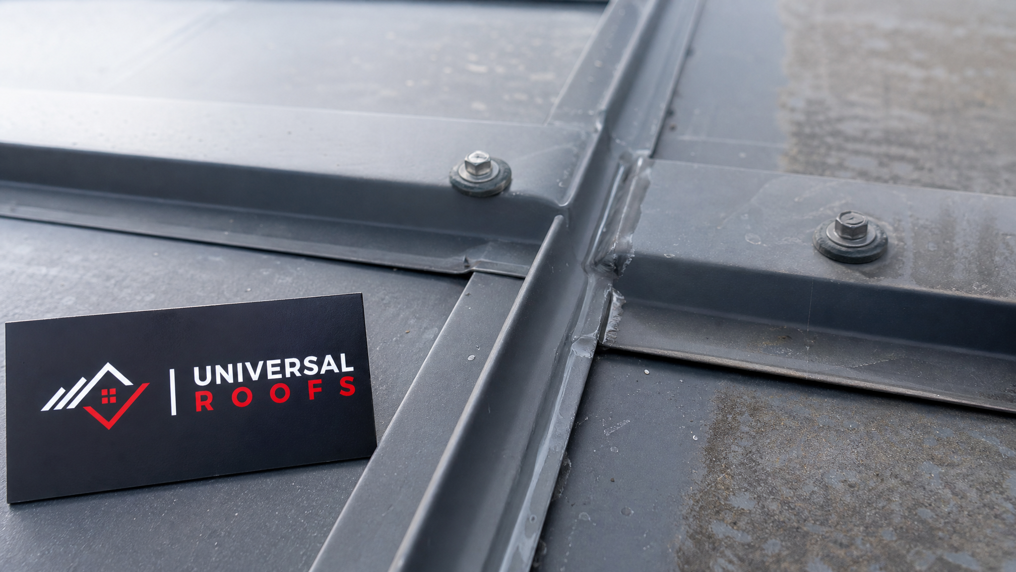Close-up detail of a repaired standing-seam metal roof panel joint with sealant and fasteners visible next to a Universal Roofs branded sign placard