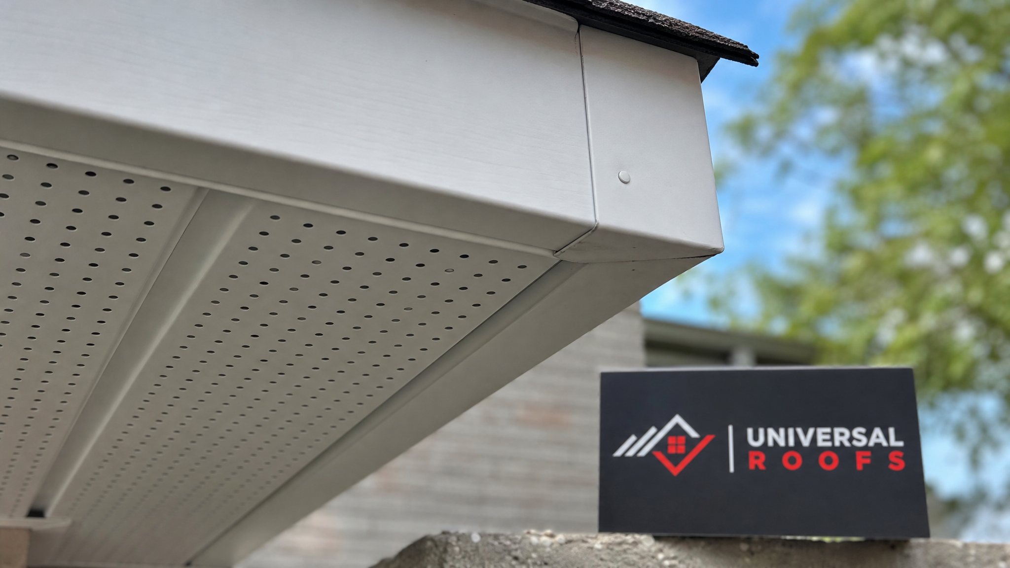 Close-up of new aluminum soffit ventilation panel and fascia board joint with a Universal Roofs branded sign placard visible
